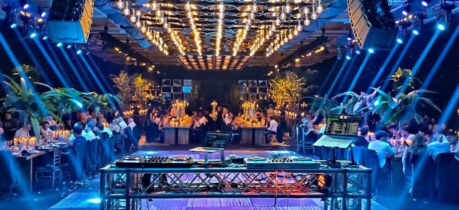 Cabina de DJ sobre estructura de truss con techo de luces cálidas y haces azules en fiesta — sonido e iluminación Stöessel